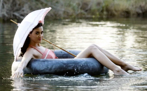 A singer with long black hair relaxes barefoot in a serene water setting, smiling while holding a pink umbrella, creating a vibrant and joyful scene.
