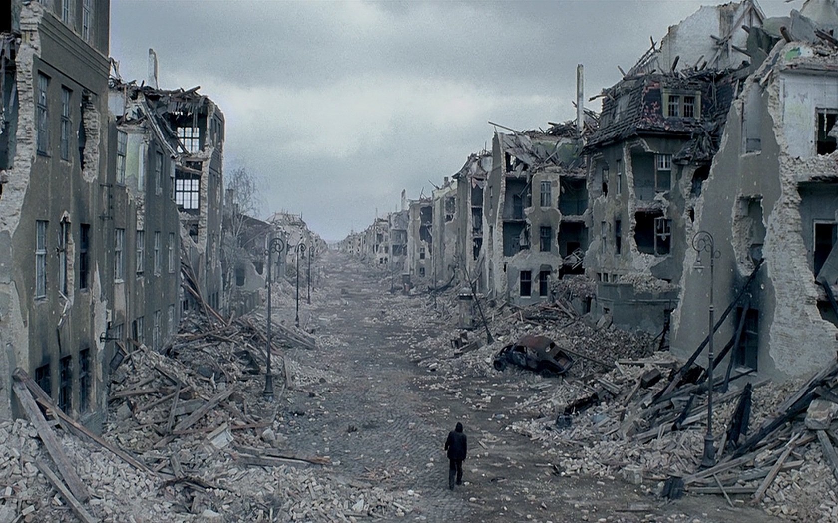 Artistic HD PC desktop wallpaper depicting a lone figure walking through a desolate, war-torn cityscape under a gray, overcast sky.
