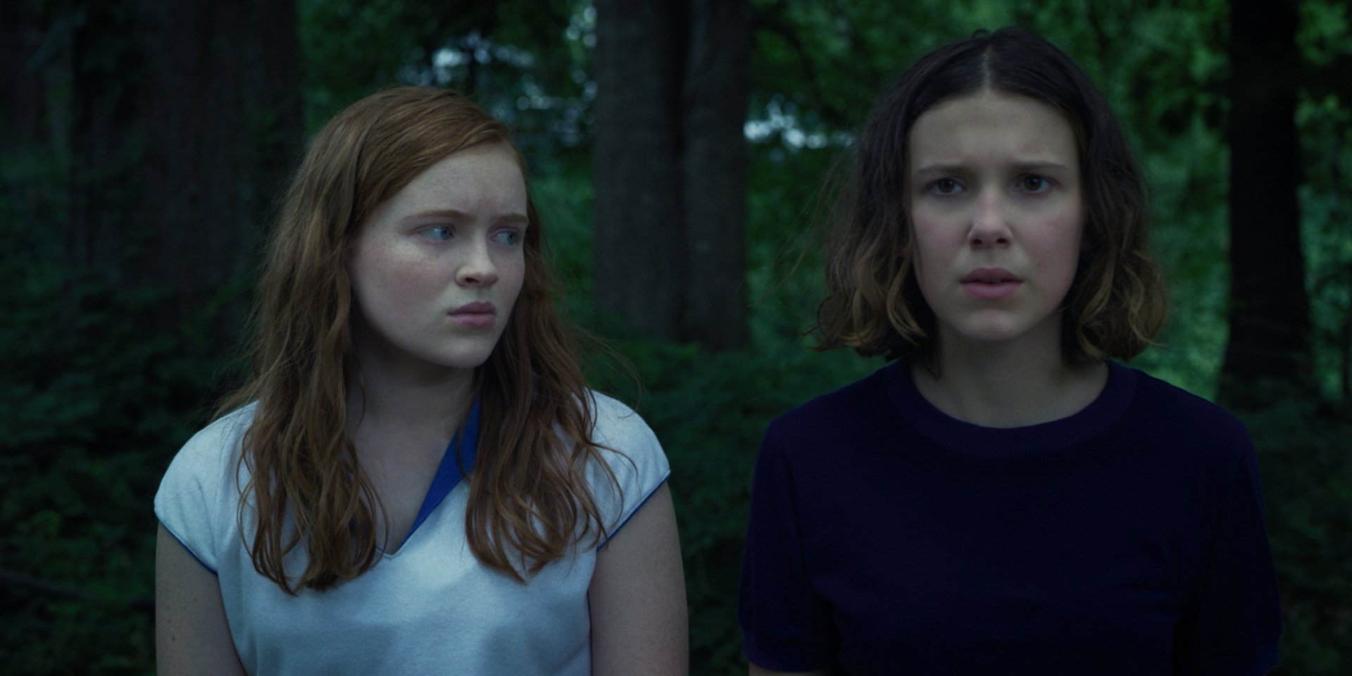 HD desktop wallpaper: two worried teenage girls in a shadowy forest with a cinematic Stranger Things TV show vibe — one redhead and one short-haired, tense expressions.