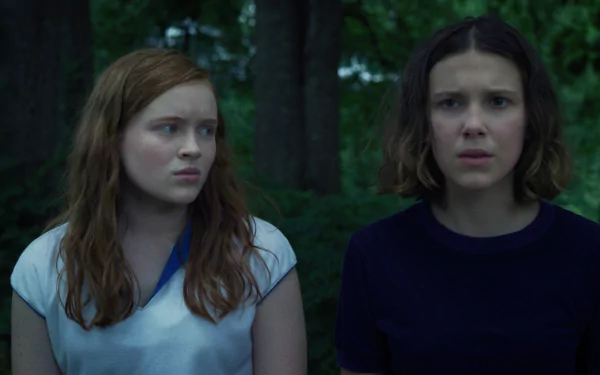 HD desktop wallpaper: two worried teenage girls in a shadowy forest with a cinematic Stranger Things TV show vibe — one redhead and one short-haired, tense expressions.