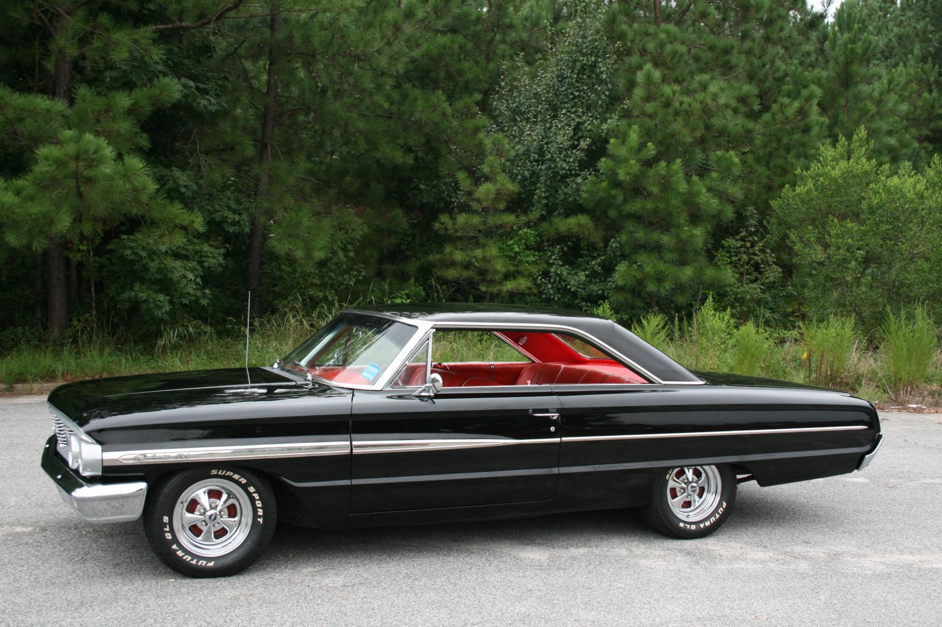 HD PC desktop wallpaper featuring a classic black Ford Galaxie 500 parked on a road with a lush green forest background.