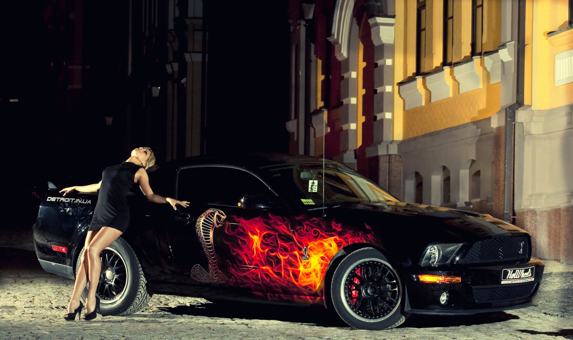 A striking HD desktop wallpaper featuring a Ford Mustang with a flaming design, complemented by a model in a stylish black dress, set against an urban backdrop.