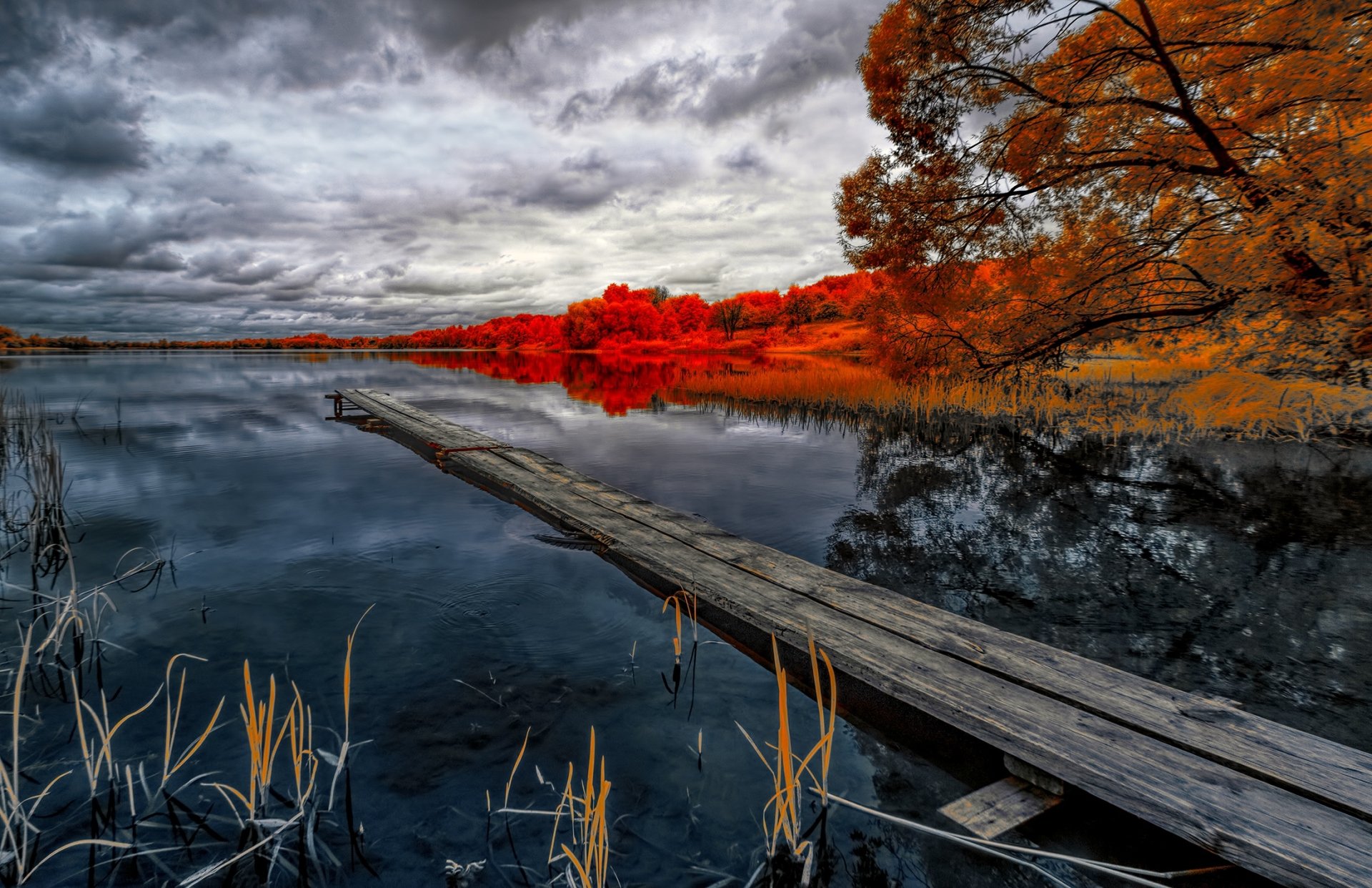 HD photography wallpaper of a calm lake with vibrant red autumn trees and a wooden dock extending into the water under a dramatic cloudy sky.