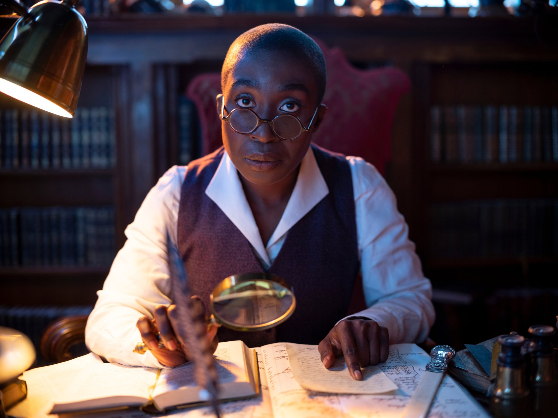 TV show The Sandman: close-up of a scholar with round glasses and magnifying glass at a desk in a moody library — 2K Quad HD PC desktop wallpaper and background.