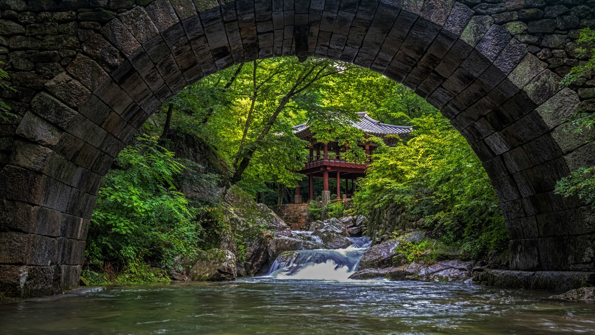 4K Ultra HD wallpaper featuring a serene religious temple nestled in lush greenery, viewed through an ancient stone arch over a flowing stream.