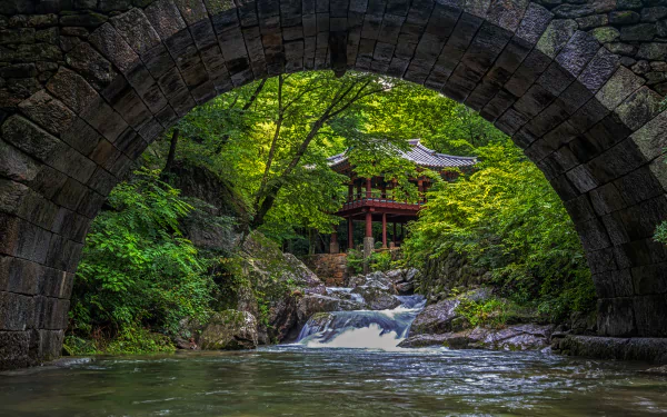 4K Ultra HD wallpaper featuring a serene religious temple nestled in lush greenery, viewed through an ancient stone arch over a flowing stream.