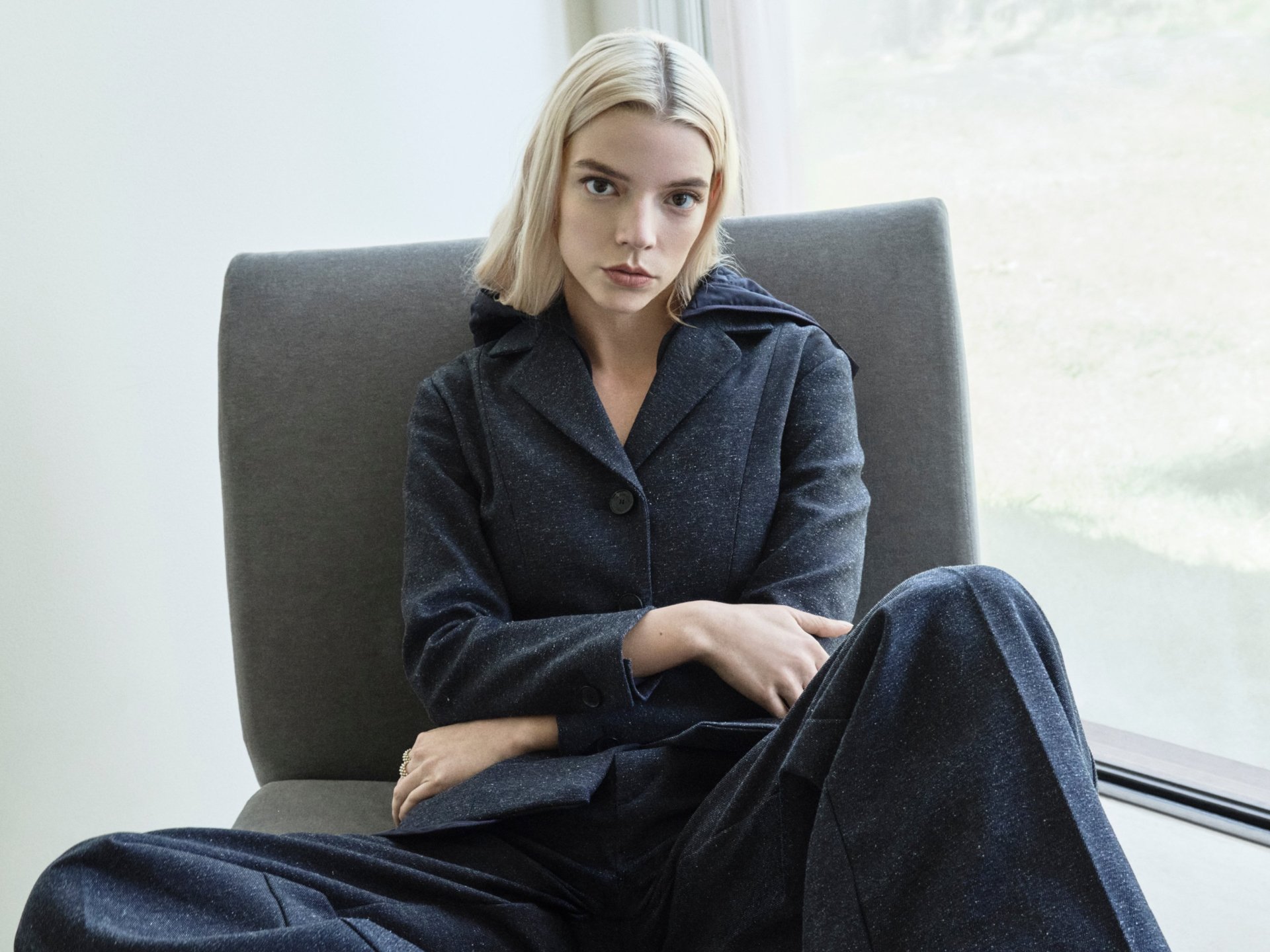 HD PC desktop wallpaper featuring celebrity Anya Taylor-Joy seated with arms crossed, dressed in dark clothing against a minimalist background.