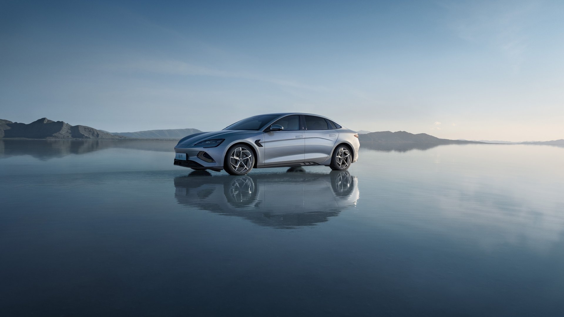 Sleek silver BYD Seal electric vehicle reflected on a glassy water surface against misty mountains — 2K Quad HD PC desktop wallpaper and background.
