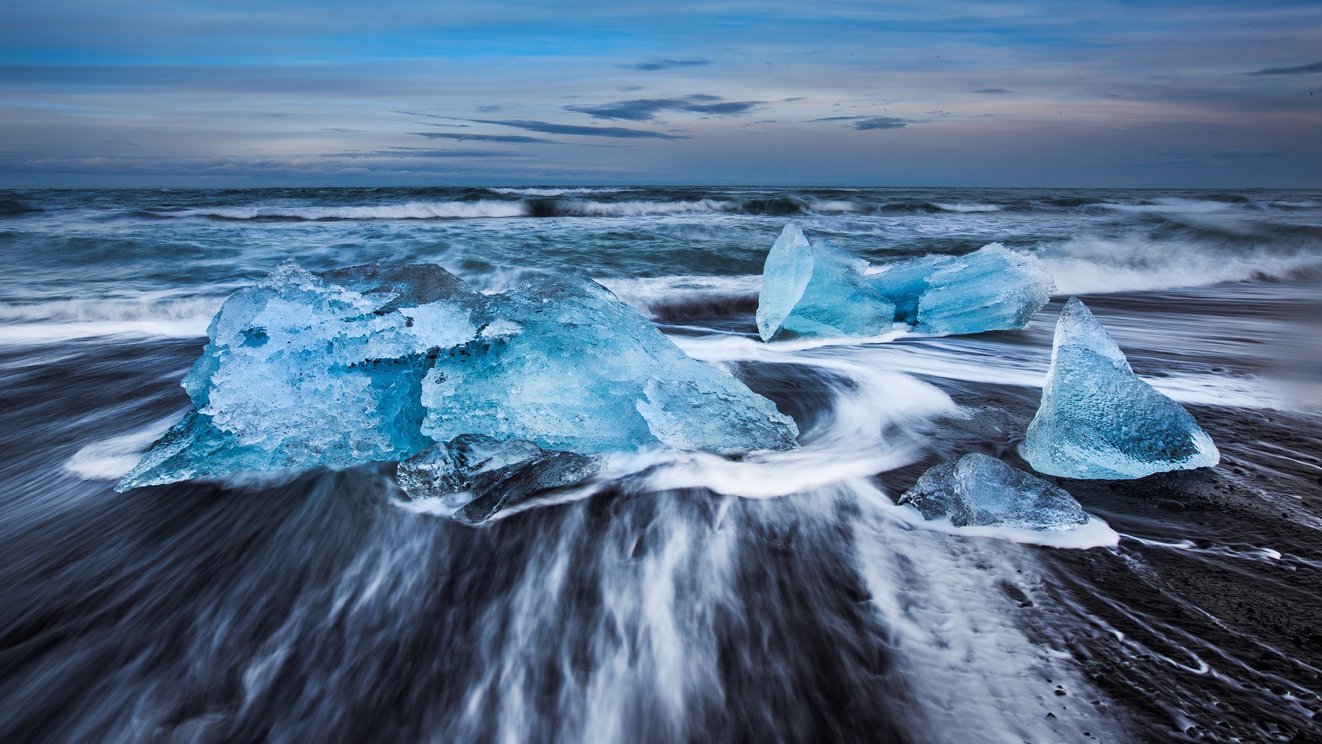 HD PC desktop wallpaper showcasing nature's beauty with translucent blue ice chunks scattered on a black sand ocean shore under a cloudy sky.