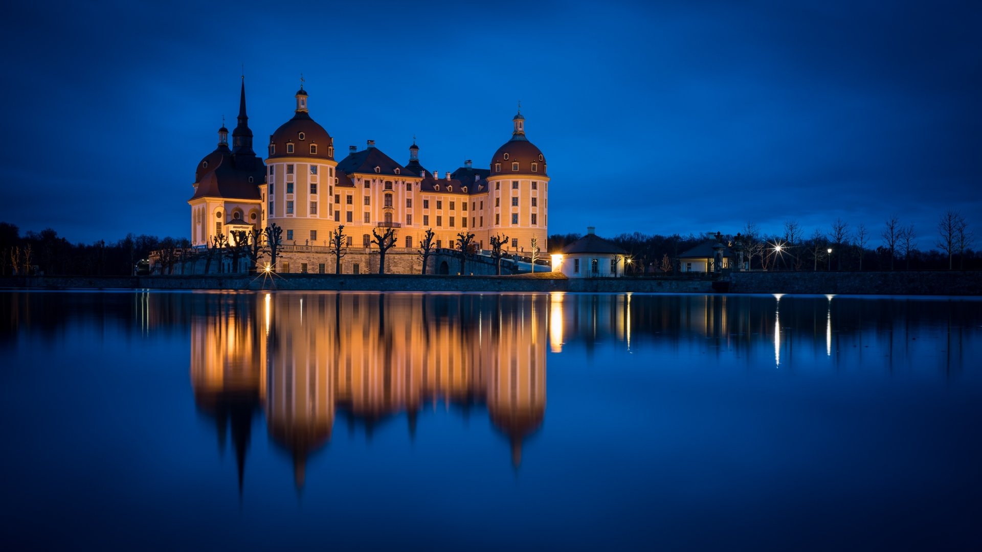 Moritzburg Castle in Germany illuminated at night, reflected perfectly in the calm water, captured as an HD desktop wallpaper showcasing man-made architectural beauty.