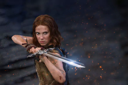 HD desktop wallpaper featuring Millie Bobby Brown as a fierce warrior from the movie Damsel, holding a sword with sparks flying in a dark, intense setting.