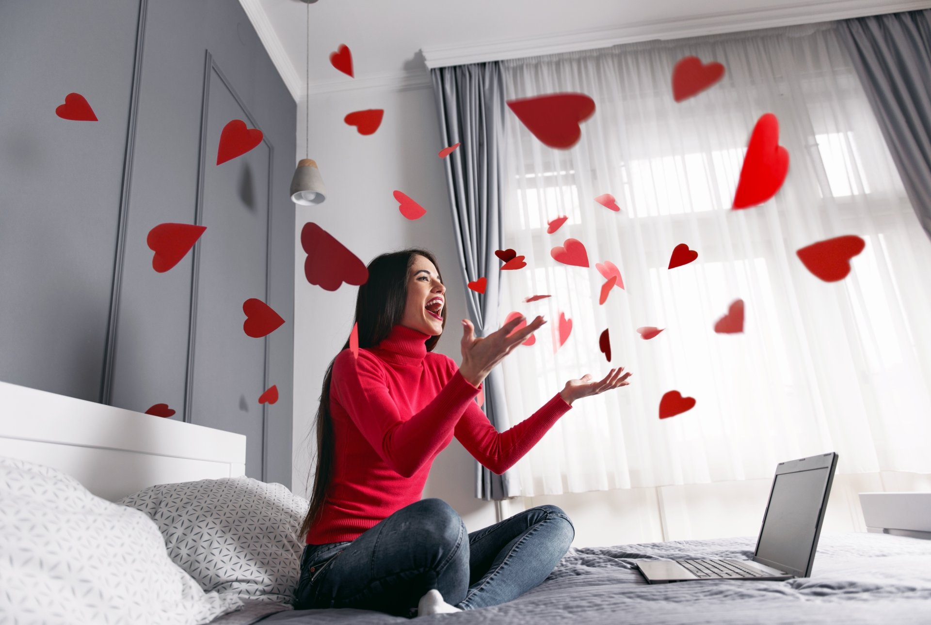 A woman in a red sweater sits on a bed, joyfully tossing red heart shapes into the air, captured in vibrant 4K Ultra HD for a PC desktop wallpaper.