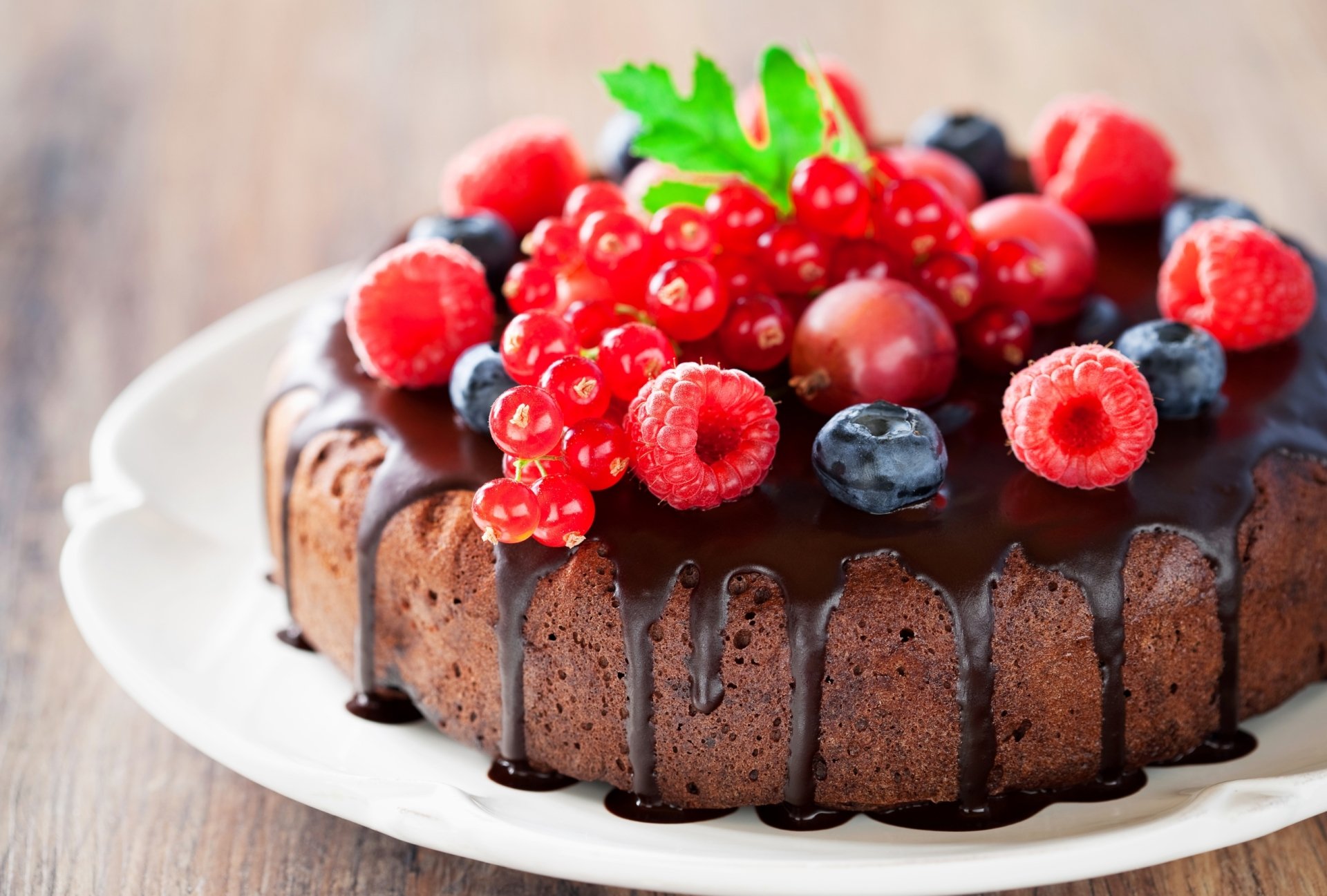 HD desktop wallpaper of a rich chocolate cake topped with mixed berries including raspberries, red currants, and blueberries on a white plate.