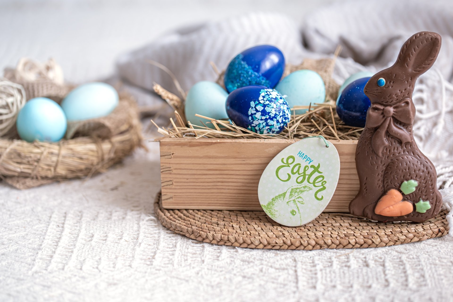 A 4K Ultra HD Easter holiday desktop wallpaper featuring colorful decorated eggs in nests and a chocolate bunny with carrots on a cozy textured surface.