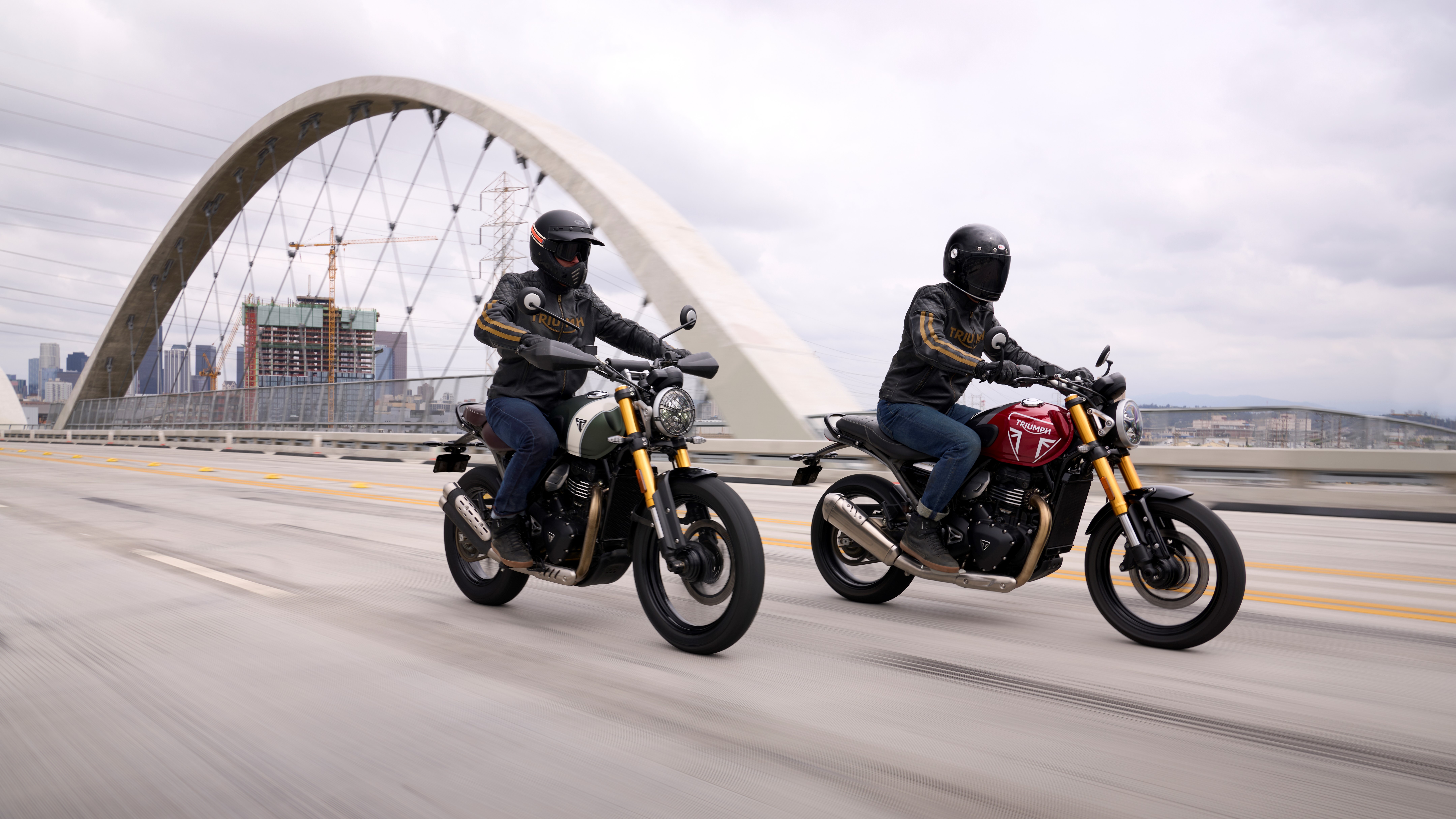 Two riders on Triumph motorcycles, with a Triumph Speed 400 and a Triumph Scrambler 400 X, cruising on a highway with an arch bridge in the background, suitable for an HD desktop wallpaper and background.