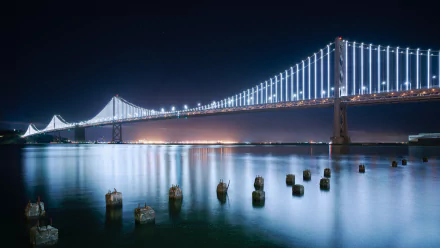 Illuminated Bay Bridge at night over calm water with reflected lights and foreground pilings — 5K Ultra HD man-made PC desktop wallpaper background.