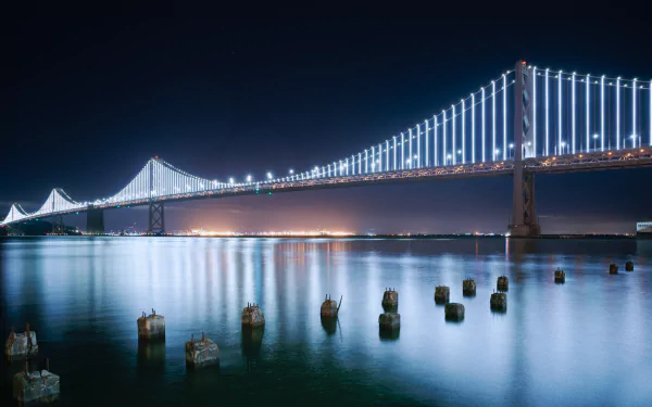 Illuminated Bay Bridge at night over calm water with reflected lights and foreground pilings — 5K Ultra HD man-made PC desktop wallpaper background.