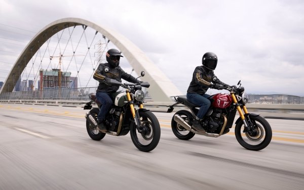 Two riders on Triumph motorcycles, with a Triumph Speed 400 and a Triumph Scrambler 400 X, cruising on a highway with an arch bridge in the background, suitable for an HD desktop wallpaper and background.