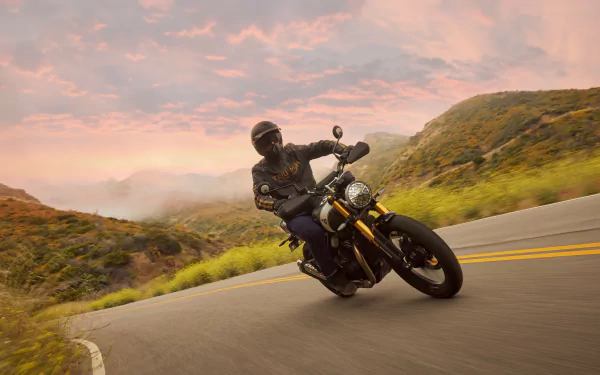 A Triumph Scrambler 400 X motorcycle leans into a curve on a scenic mountain road, captured in a stunning HD wallpaper with a vibrant sunset backdrop.