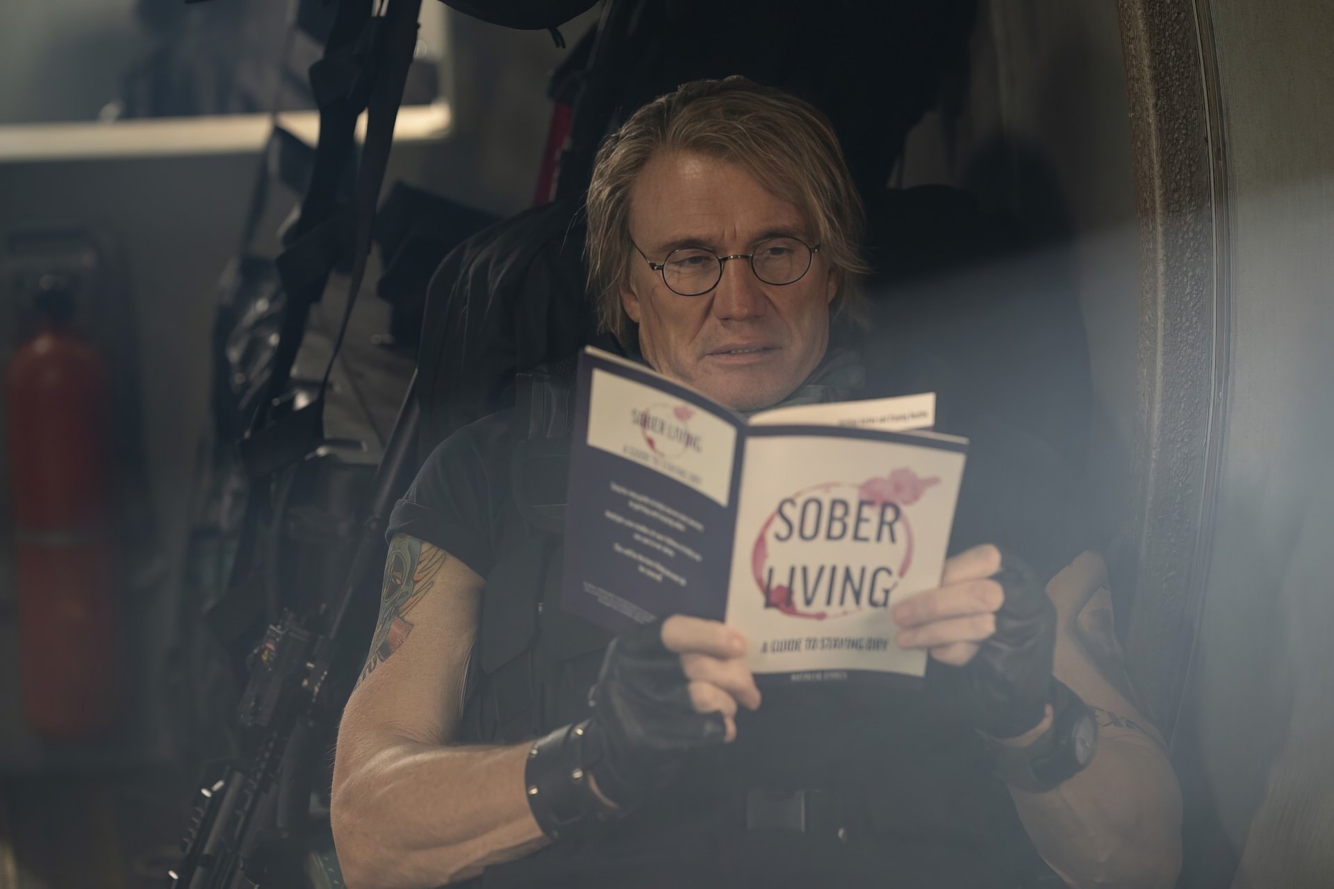 HD wallpaper featuring a person with long blonde hair and glasses, reading a book titled 'SOBER LIVING' in a dimly lit room, with elements related to the Expendables theme visible in the background.