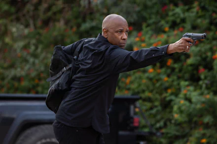 An intense scene from The Equalizer 3, featuring a man in a black outfit aiming a gun, set against a backdrop of green foliage and vibrant flowers. HD wallpaper option.