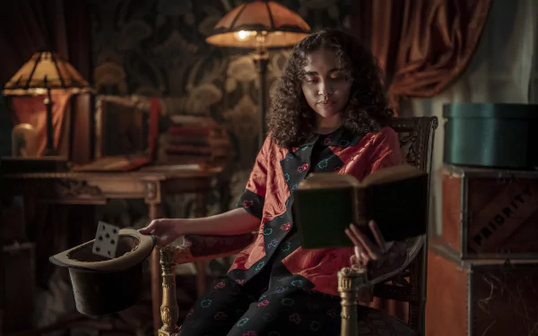 Bailey Bass sits thoughtfully in a cozy, vintage setting, reading a book while holding a top hat, showcasing a scene from the TV show Interview with the Vampire in high-definition detail.