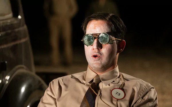 HD desktop wallpaper featuring a man in a vintage tan suit and sunglasses with an Oppenheimer tag, evoking a historical theme against a blurred background.