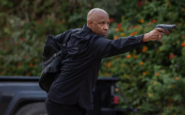 An intense scene from The Equalizer 3, featuring a man in a black outfit aiming a gun, set against a backdrop of green foliage and vibrant flowers. HD wallpaper option.