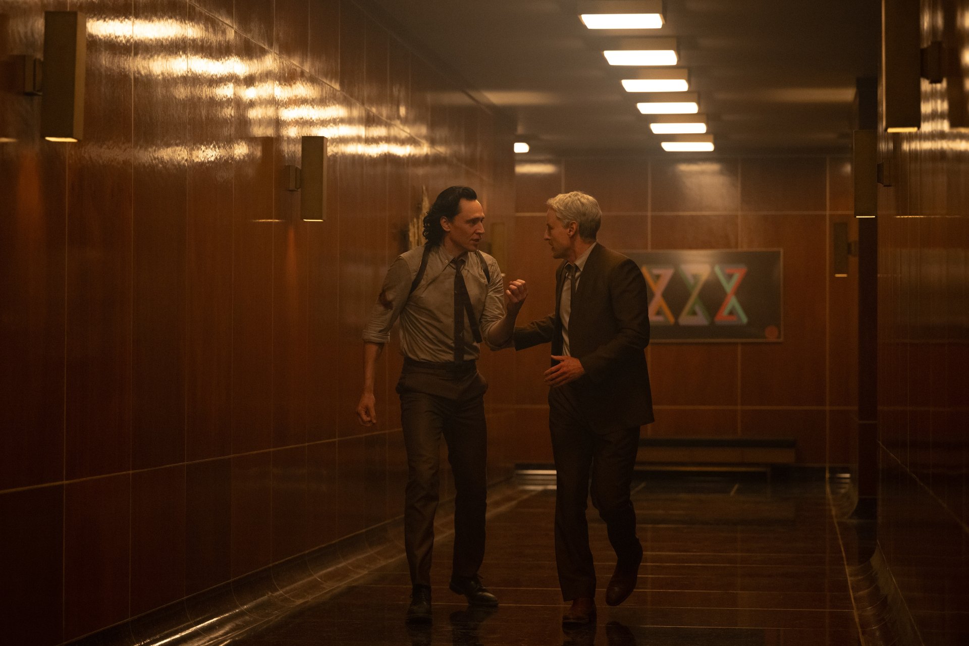 HD desktop wallpaper featuring two characters in a conversation from the Loki series, set in a moody, dimly lit corridor with futuristic symbols on the wall.