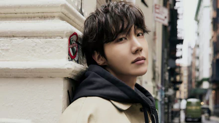 A high-definition desktop wallpaper featuring J-Hope, a member of BTS, leaning against a wall with a contemplative expression, surrounded by an urban backdrop.