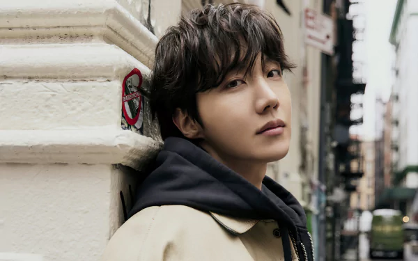 A high-definition desktop wallpaper featuring J-Hope, a member of BTS, leaning against a wall with a contemplative expression, surrounded by an urban backdrop.