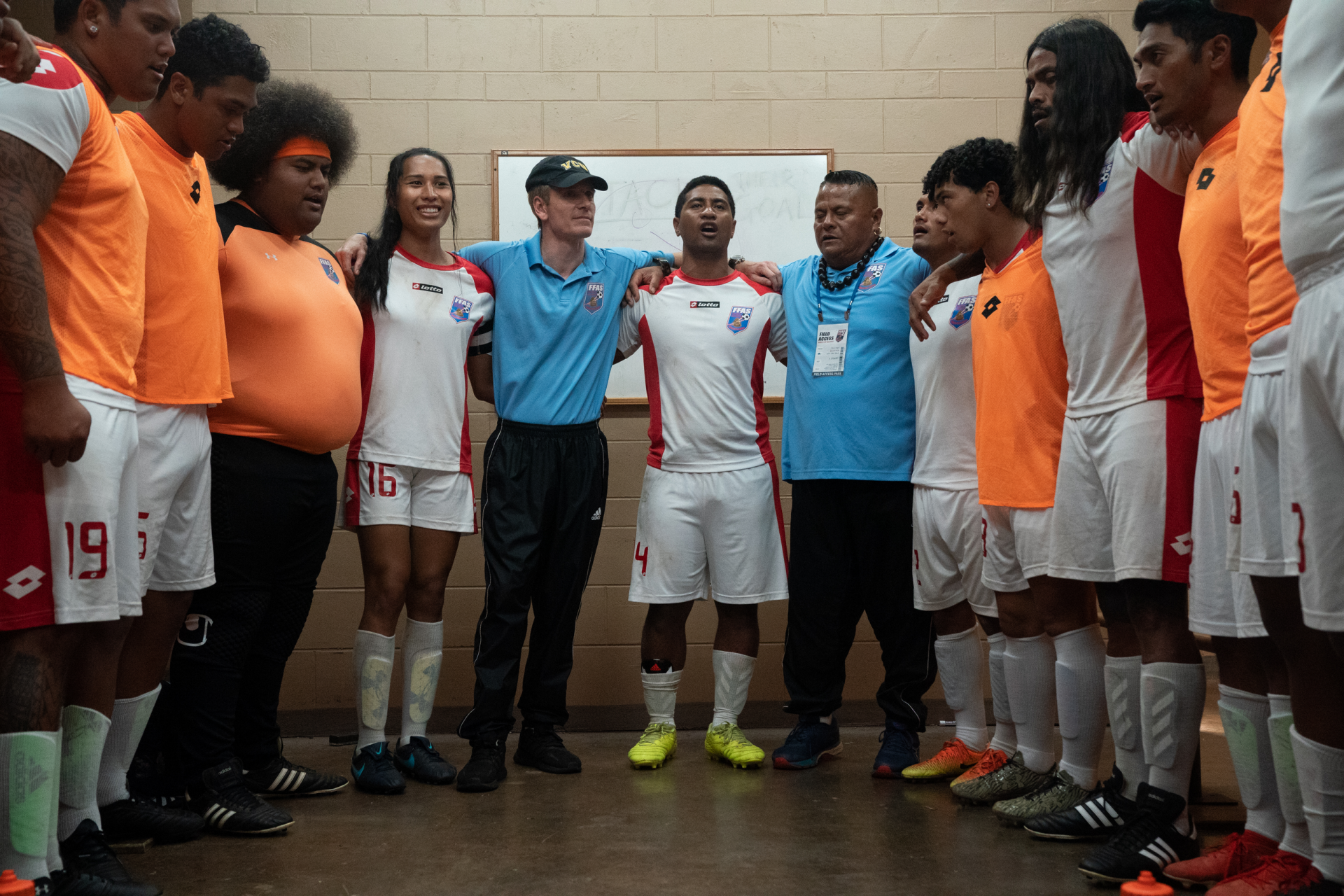 A scene from the movie *Next Goal Wins* (2023), featuring a diverse soccer team in a locker room, united in a circle before a match, showcasing camaraderie and determination.