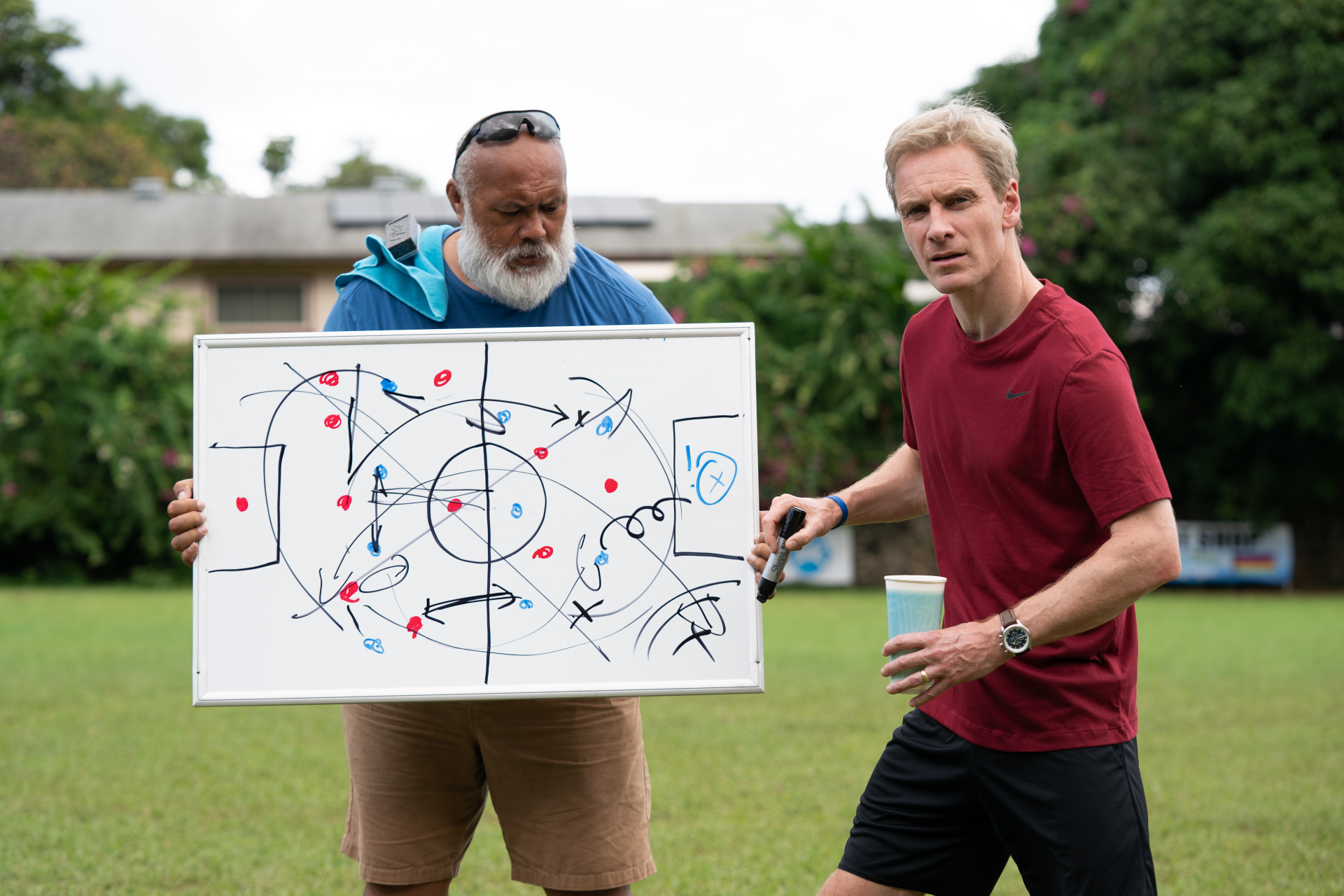 Two male characters from the movie Next Goal Wins (2023) discuss strategy using a soccer playbook in an HD desktop wallpaper.