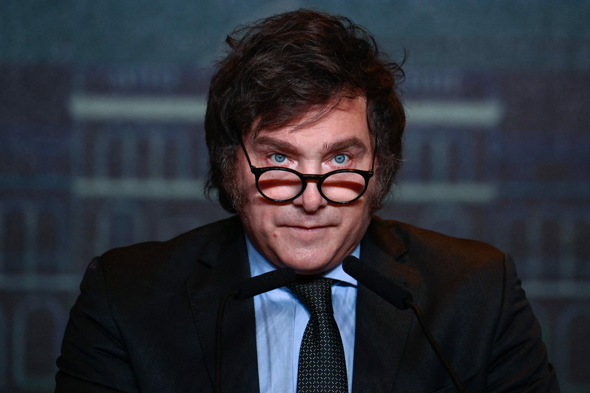 HD desktop wallpaper of a person with unique glasses, a stern expression, dressed in a suit, with a blurred background.