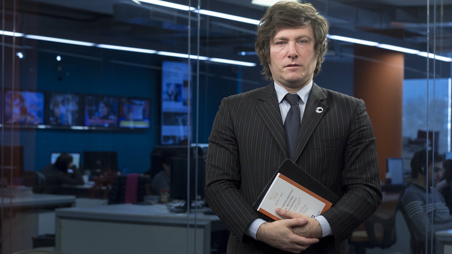 Elegant man in a suit holding a book with a newsroom background for HD desktop wallpaper.