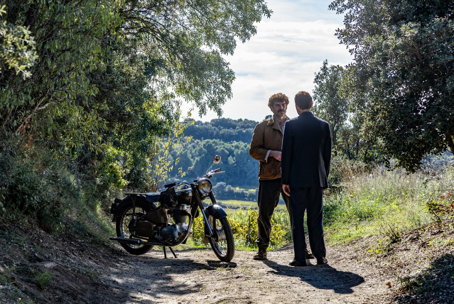 HD wallpaper of two characters from the TV show 'Monsieur Spade' chatting beside a motorcycle on a picturesque countryside path.