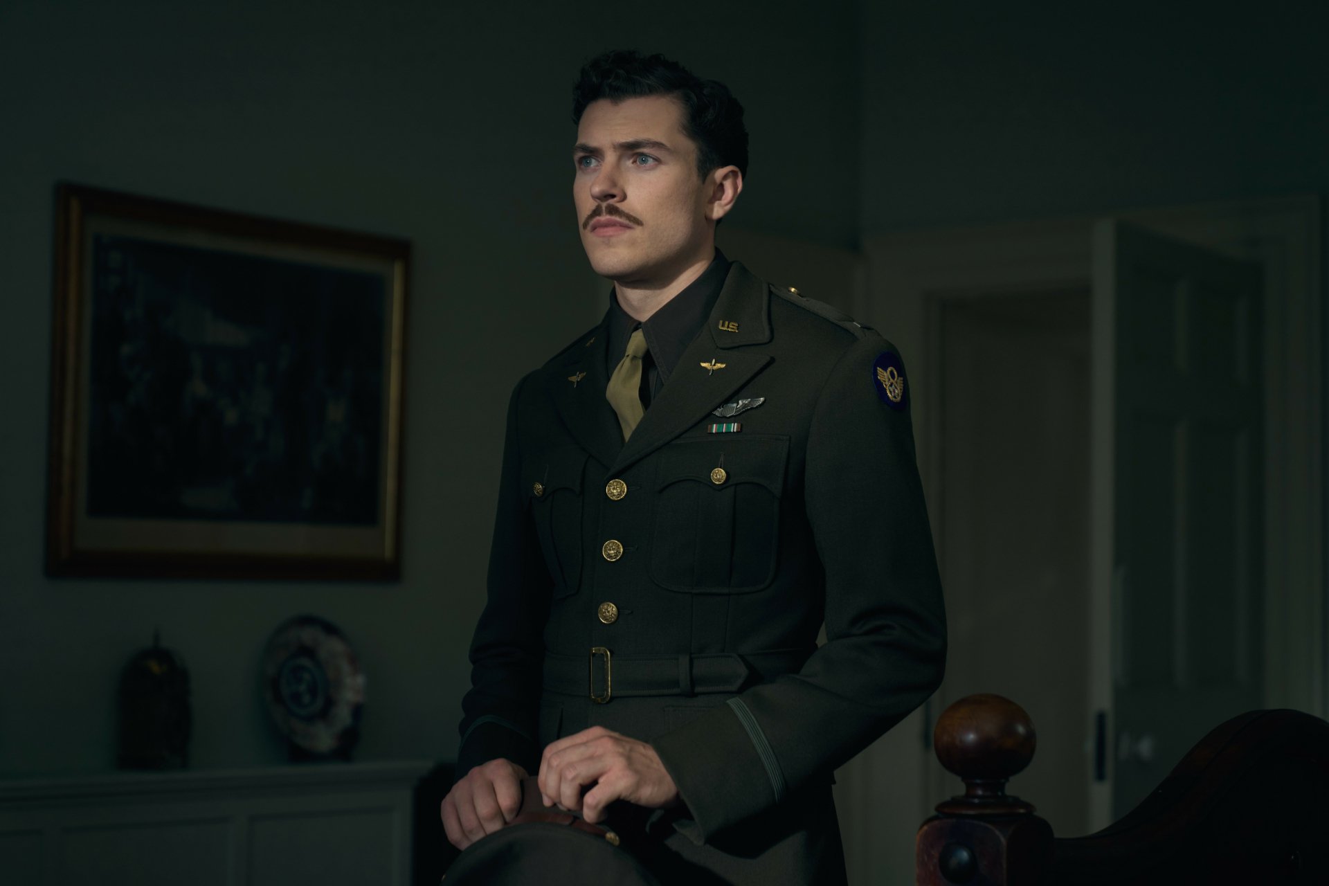 HD wallpaper of an intense scene from the TV show Masters of the Air featuring a military officer in uniform standing thoughtfully in a dimly lit room.