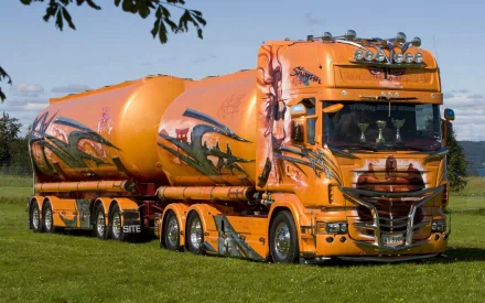 A vibrant orange Scania truck with striking artwork, set against a grassy background under a clear blue sky. This HD image serves as an eye-catching desktop wallpaper.