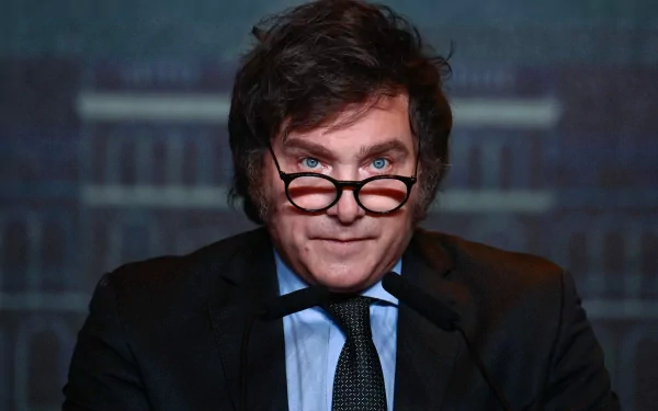 HD desktop wallpaper of a person with unique glasses, a stern expression, dressed in a suit, with a blurred background.