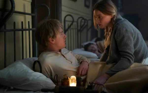 A scene from the TV show All the Light We Cannot See, featuring two characters in bed, engaged in an intense conversation, with a soft glow illuminating their faces. HD wallpaper background.