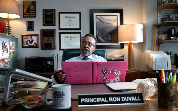 HD desktop wallpaper featuring a scene from Mean Girls (2024) with a character seated at an office desk, examining a pink binder labeled Burn Book, set in a well-decorated office displaying the nameplate PRINCIPAL RON DUVALL.