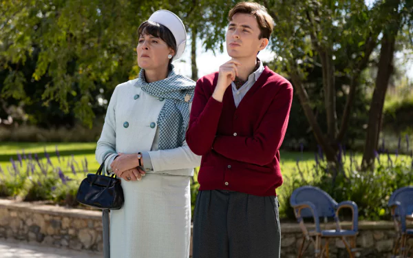 A scene from the TV show Monsieur Spade, featuring two characters in stylish attire, standing outdoors among greenery, conveying intrigue and contemplation. HD desktop wallpaper.