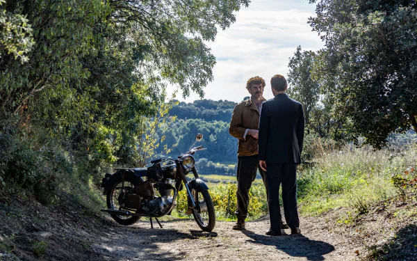 HD wallpaper of two characters from the TV show 'Monsieur Spade' chatting beside a motorcycle on a picturesque countryside path.