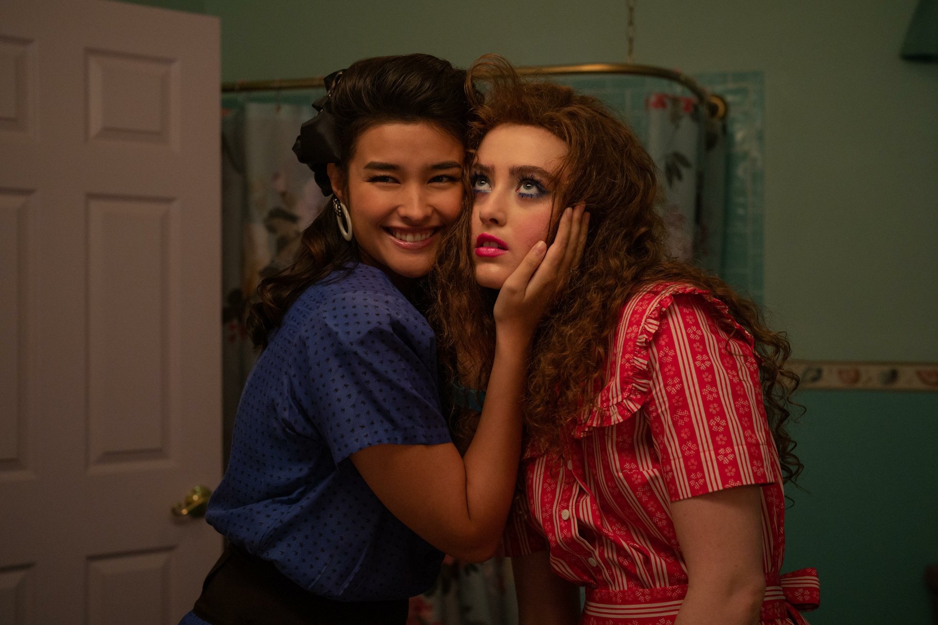 Two young women share a playful moment in a bathroom, showcasing 1950s-inspired fashion. This HD wallpaper features a scene from the movie Lisa Frankenstein.
