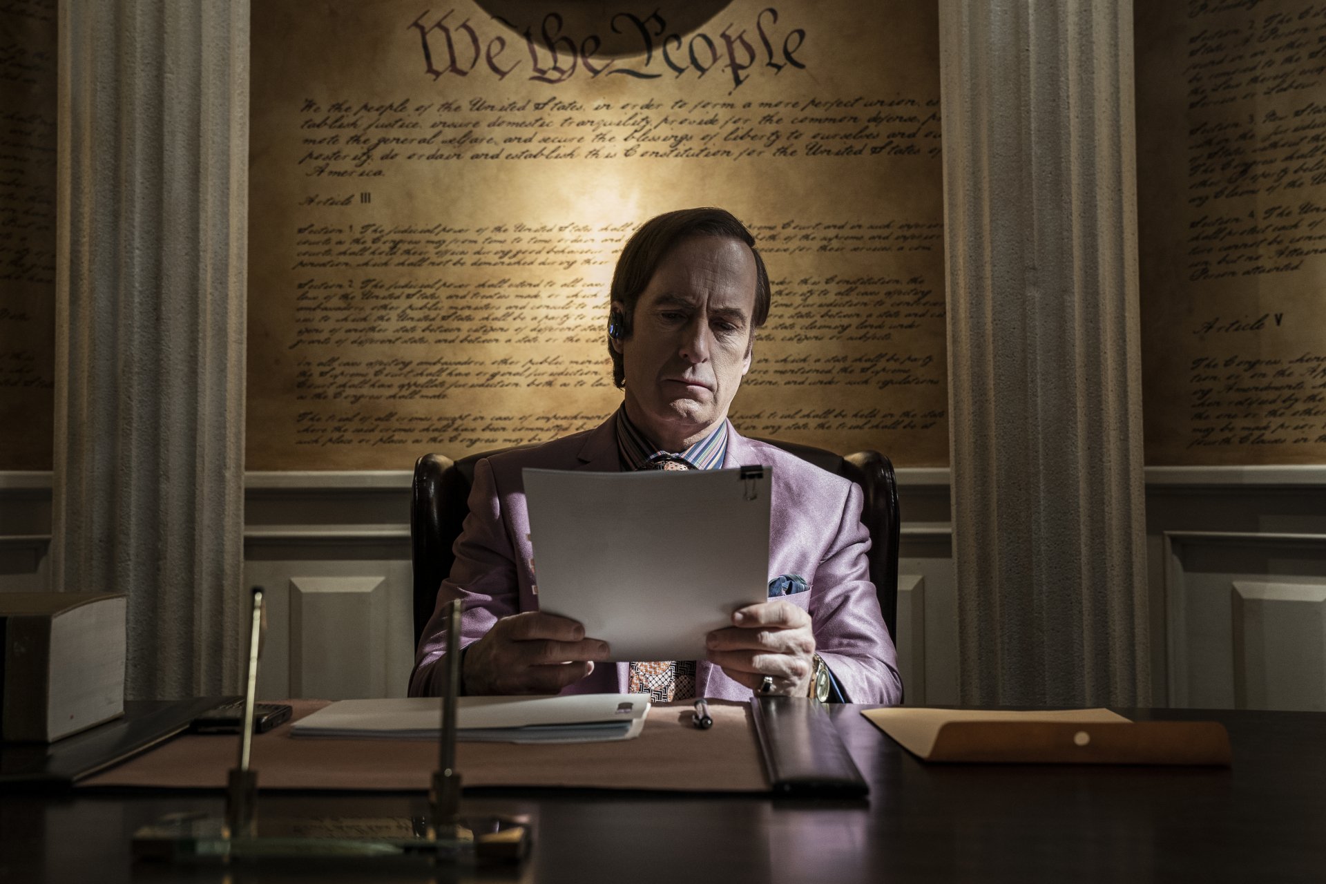 HD wallpaper featuring a man portraying a lawyer, resembling a character from the TV show Better Call Saul, sitting at a desk with a serious expression, reviewing a document, with the phrase We the People visible in the background.