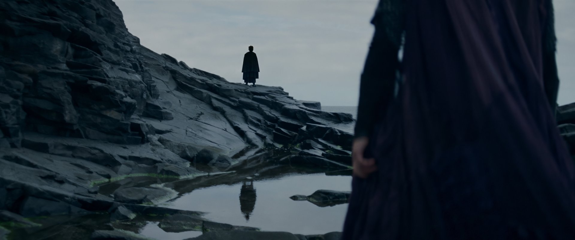 HD wallpaper featuring a scene from the TV show The Acolyte (2024) with a mysterious figure standing on a rocky landscape reflected in water.