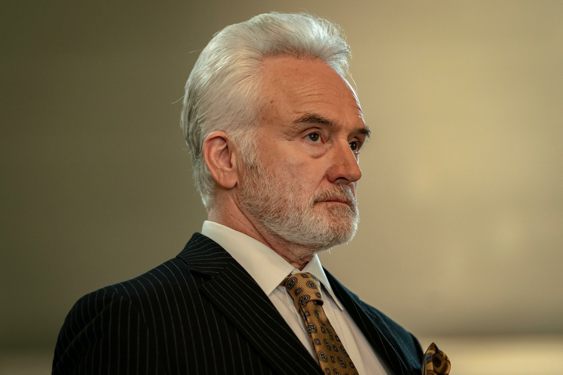 A character from the TV show Parish (2024) is portrayed with a serious expression, wearing a dark pinstripe suit and a patterned tie, set against a softly blurred background.