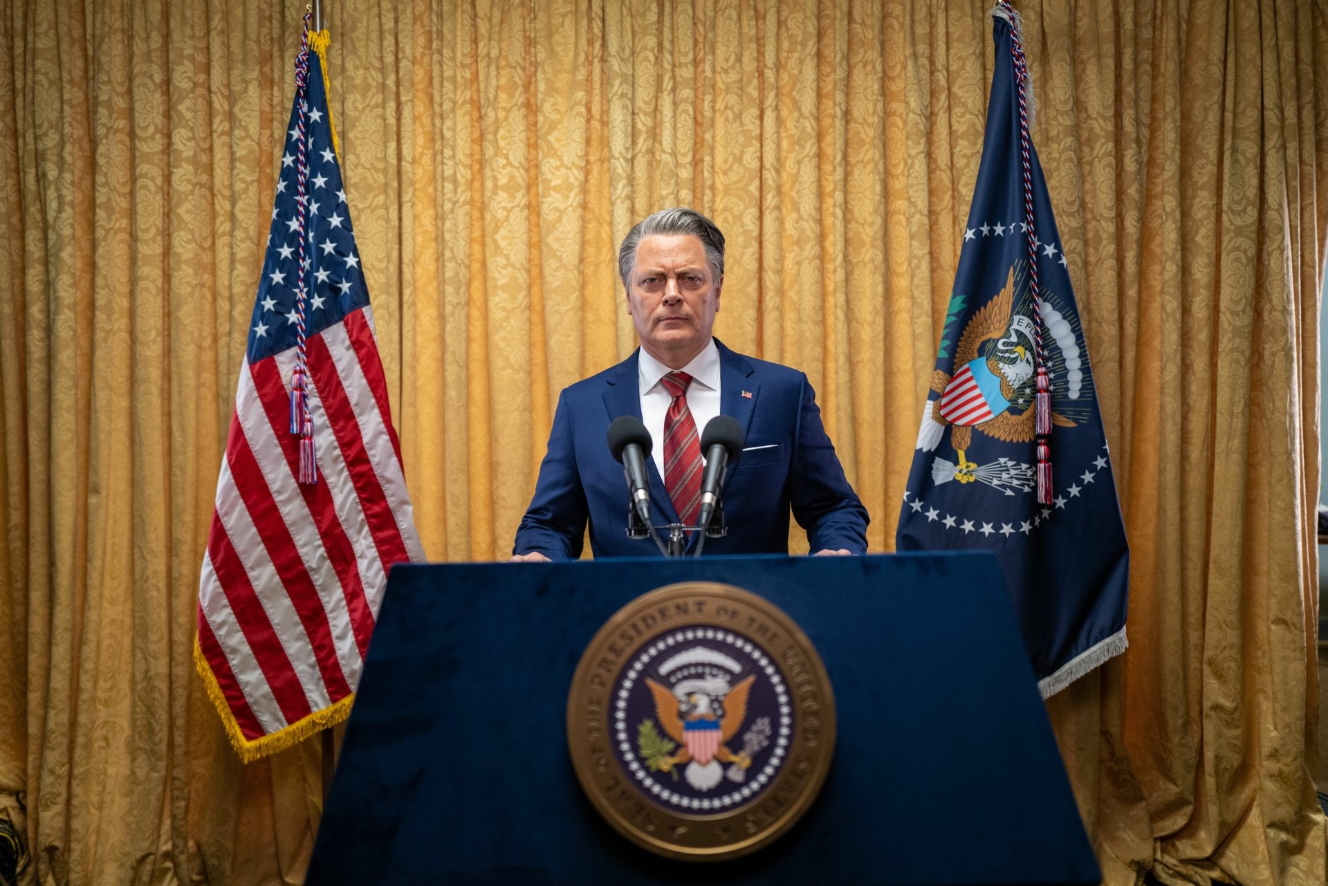 A man standing at a podium with the Presidential Seal, flanked by American flags, in a scene from the movie Civil War (2024)—an HD desktop wallpaper background.