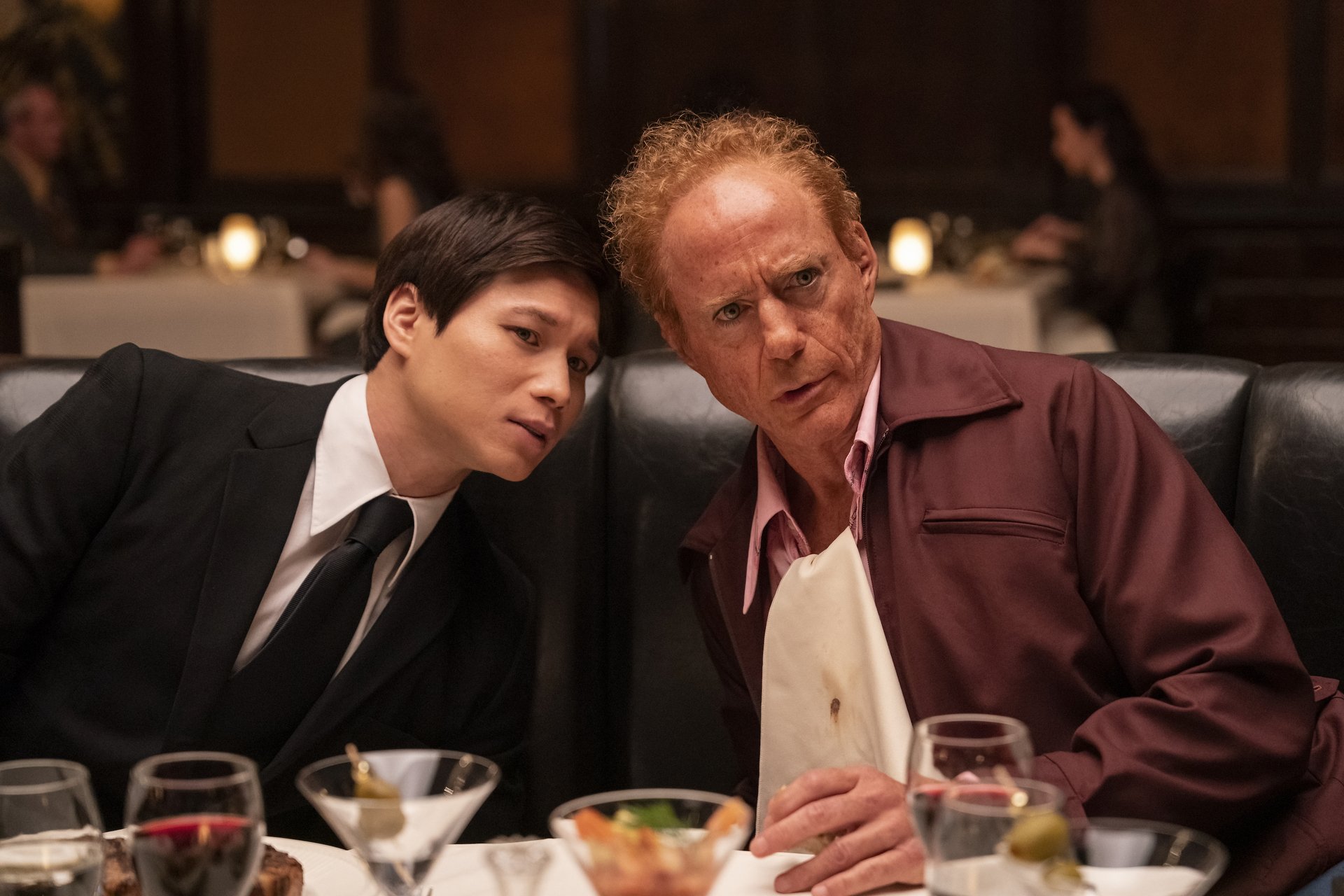 HD wallpaper featuring two characters from the TV show The Sympathizer, a young man in a suit and an older man in a maroon shirt, leaning closely together at a restaurant, possibly engaged in an intense conversation.