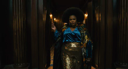 A striking scene from the TV show The American Society of Magical Negroes, featuring a confident figure in a vibrant outfit, set in a dimly lit corridor adorned with warm lighting.